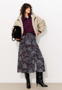 Doudoune beige, haut zippé violet foncé, jupe à motifs floraux en bordeaux et bleu marine, bottes noires à talons hauts jusqu'aux genoux, portant un sac noir.