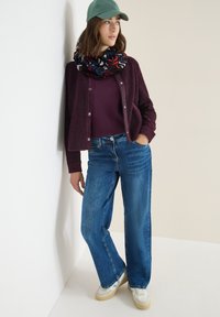 Burgundy fuzzy cardigan, dark purple top, blue wide-leg jeans, patterned black scarf, and green cap paired with cream sneakers.
