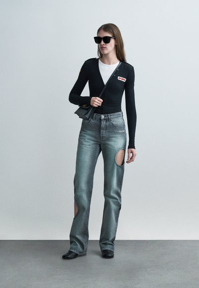 Femme portant des lunettes de soleil noires, un cardigan noir ajusté sur un haut blanc, un jean délavé avec de grandes découpes et des chaussures noires, tenant un petit sac noir.