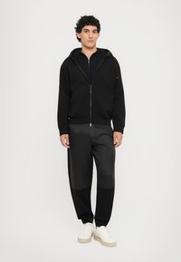 Svart zip-up hettegenser med lommer, kombinert med svarte bukser med jevn tekstur og kontrasterende paneler. Hvite joggesko fullfører looken.