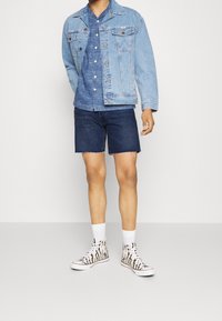 Ljusblå denimjacka, lager på lager över en mörkblå randig skjorta. Bärs med mörk denimshorts och vita strumpor ihop med hög-top snakers i zebramönster.