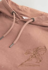 Sweat à capuche rose poussiéreux avec cordons tressés et embouts en métal, orné d'une broderie florale discrète sur la poitrine.
