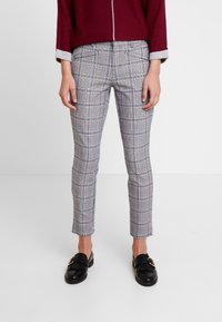 Personne portant un pantalon slim en plaid gris et des mocassins noirs, avec un haut à manches longues bordeaux partiellement visible, se tenant sur un sol blanc.