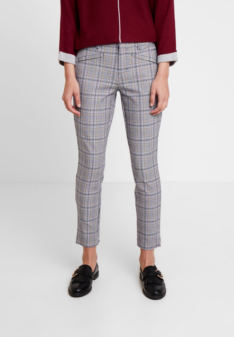 Personne portant un pantalon slim en plaid gris et des mocassins noirs, avec un haut à manches longues bordeaux partiellement visible, se tenant sur un sol blanc.