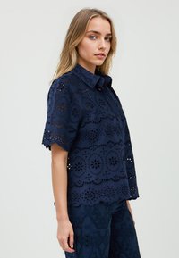 Giovane donna con lunghi capelli mossi posa di lato, indossando una camicia ricamata blu navy con motivi floreali su uno sfondo neutro.