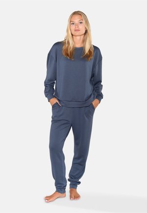 Lyshåret kvinde står barfodet iført en løs mørkeblå sweatshirt og matchende joggerbukser med hænderne i lommerne mod en ensfarvet baggrund.