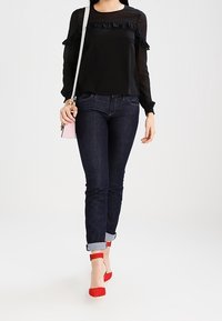 Sort bluse med lange ærmer og flæse-detaljer, parret med mørke denimjeans. Røde ankelrem sandaler og en lille lyserød crossbody taske.