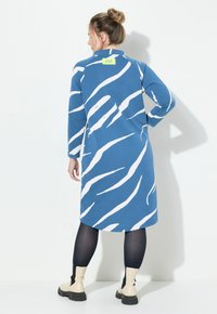 Blauwe jurk met witte abstracte strepen, voorzien van een groene label aan de kraag. Lange mouwen, halflengte, gedragen met zwarte legging en beige laarzen.