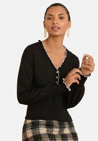 Schwarzer Pullover mit weißen Rüschendetails, V-Ausschnitt und Knopfdetails. Das Model trägt ihn über einem karierten Rock, der ein figurnahes Design und strukturierten Stoff zeigt.