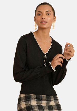 Maglione nero con dettagli in pizzo bianco, scollatura a V e dettagli con bottoni. Il modello lo indossa su una gonna a quadri, mettendo in risalto un design aderente e un tessuto texture.