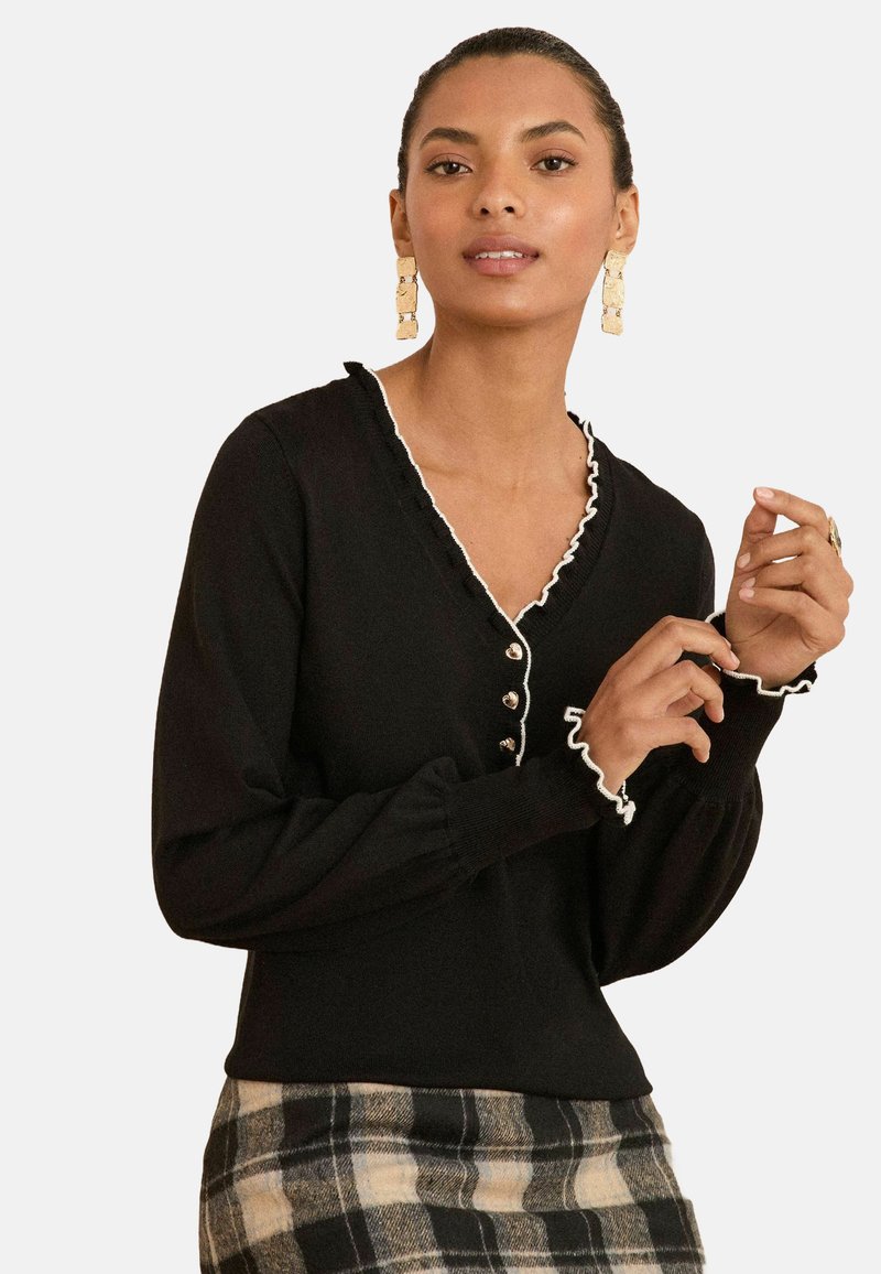 Schwarzer Pullover mit weißen Rüschendetails, V-Ausschnitt und Knopfdetails. Das Model trägt ihn über einem karierten Rock, der ein figurnahes Design und strukturierten Stoff zeigt.