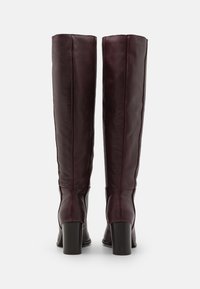 Bottes en cuir bordeaux montantes avec une texture lisse, bout rond et solide talon bloc. Détails de couture visibles sur la tige.