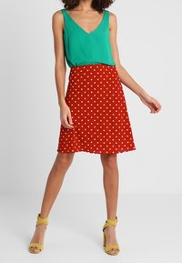 Zielony bezrękawnik, czerwona spódnica w kropki z falbaniastym dołem oraz żółte sandały na wysokim obcasie. Gładka faktura i żywe kolory.
