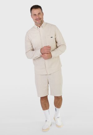 Lächelnder Mann steht vor einfarbigem Hintergrund, trägt ein beiges Hemd mit Brusttasche, passende Shorts, weiße hohe Socken und weiße Sneakers.