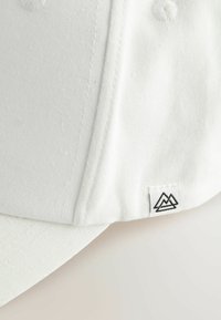 Hvid bomuldsbaseballkasket med en struktureret design, der har en flad skygge. Inkluderer en lille sort bjerglogoetiket på siden.