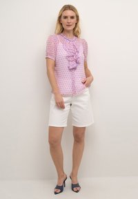 Rosa Bluse mit Diamantmuster, Rüschendetail entlang der Knopfleiste, kurze Ärmel. Kombiniert mit weißen Shorts und blauen Absatzschuhen.