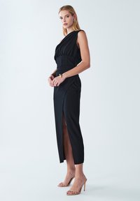 Abito nero con corpino aderente e scollo drappeggiato. Presenta una spacco laterale e un design affusolato. La modella indossa sandali con tacco nudo.