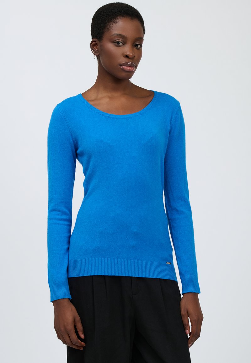 Maglione blu a maniche lunghe realizzato in tessuto morbido, con scollatura tonda e orlo a coste. Il modello lo indossa con pantaloni neri.