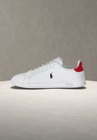 Λευκό sneaker χαμηλού κόστους με δέσιμο με κορδόνια μπροστά, μαύρο λογότυπο αλόγου του polo στο πλευρό και κόκκινη ταμπέλα στη φτέρνα με την επιγραφή «Polo», εμφανίζεται σε ουδέτερη επιφάνεια.