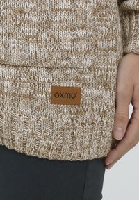 Tan ir kremas megztinis su aksominiu apvadu ir rudos odos etikete su "OXMO" logotipu, pateikta su tamsiomis kelnėmis.