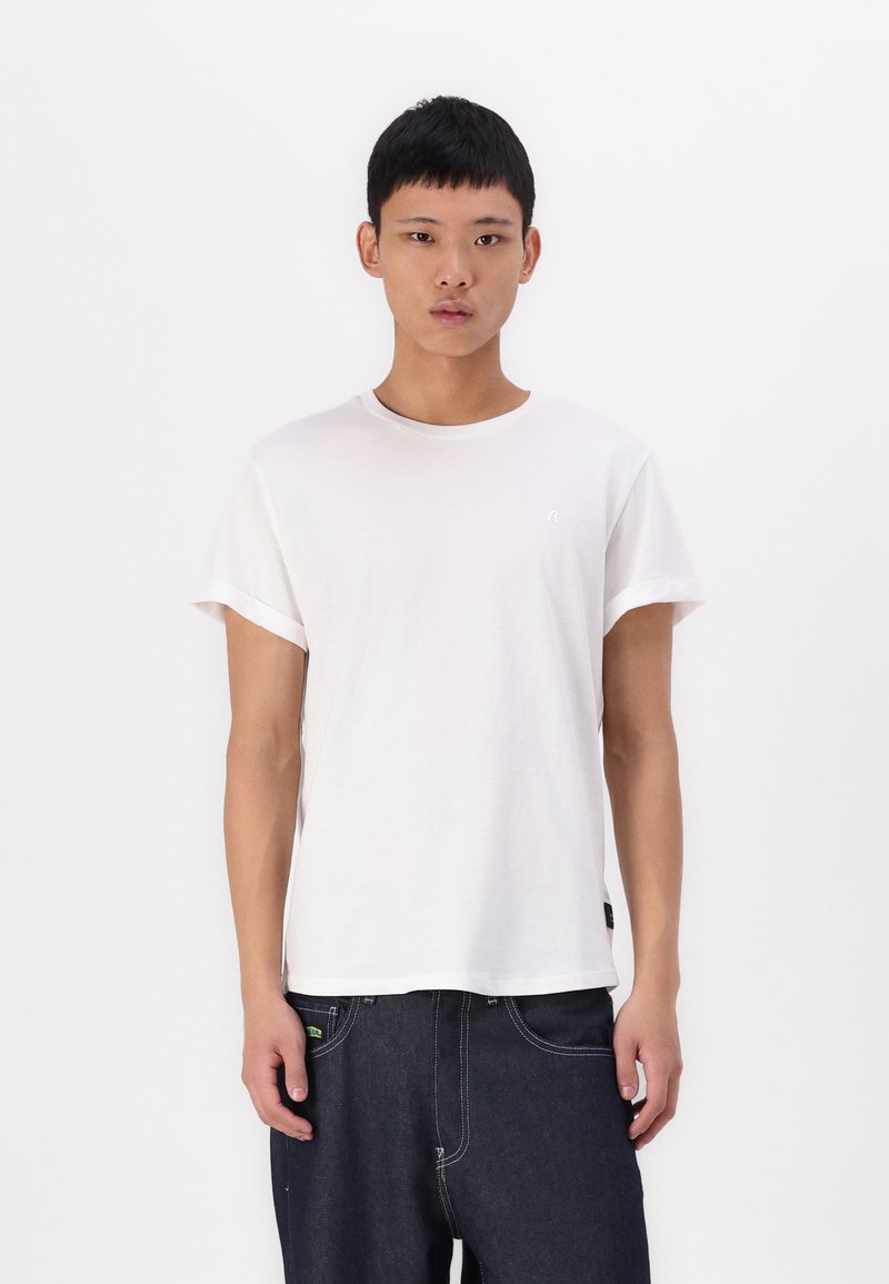Weißes Baumwoll-T-Shirt mit kurzen Ärmeln, rundem Ausschnitt und kleinem Logo auf der Brust. Kombiniert mit dunklen Jeans.