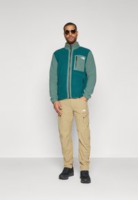 Teal- og grå fleecejakke med glidelåslommer, parret med beige cargobukser og svarte sko. Modell iført solbriller.