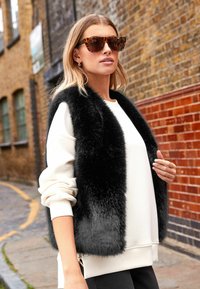 Gilet in pelliccia sintetica nera sopra un sweatshirt bianco, abbinato a occhiali da sole tartaruga. Presenta una zip all'orlo, con una finitura morbida e testurizzata.