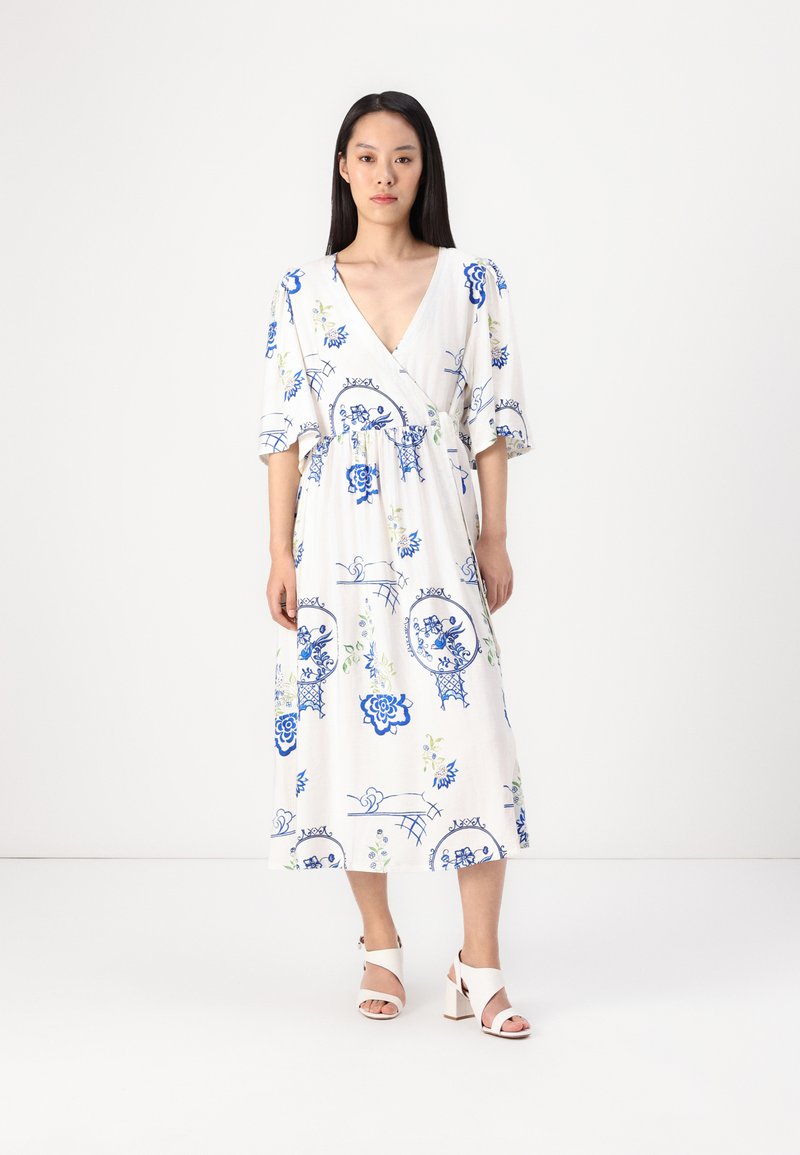 Weißes Midi-Kleid mit blauem Blumendruck, tiefem V-Ausschnitt, kurzen Puffärmeln und gerafftem Bund; kombiniert mit weißen Blockabsatzsandalen.