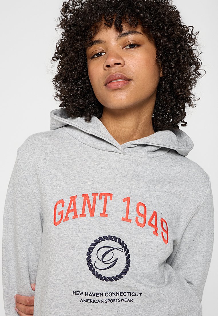 Grauer Hoodie mit rotem Text "GANT 1949" und einem runden Logo. Weicher Stoff, Kapuze mit Kordelzug und gerippte Bündchen. Einfaches, sportliches Design.