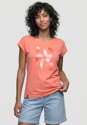 Frau mit lockigem dunklem Haar trägt ein korallenfarbenes T-Shirt mit geometrischem "LOVE"-Druck und hellblaue hochgekrempelte Shorts vor schlichtem grauem Hintergrund.