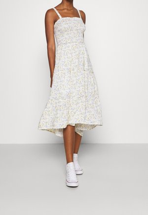 Mujer con un vestido midi blanco sin mangas con estampado floral, bajo con volantes y zapatillas blancas altas, de pie sobre un fondo liso y claro.