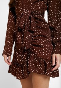 Ruskea puolipisteinen mekko, jossa on pitkät hihat, kiristetty vyötärö, röyhelöhame ja solmittava vyökoriste. Kangas näyttää pehmeältä ja teksturoidulta.