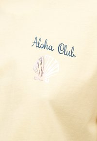 Ύφασμα σε κρεμ χρώμα με σκούρο μπλε κείμενο "Aloha Club" πάνω από κεντημένο σχέδιο κοχυλιού.