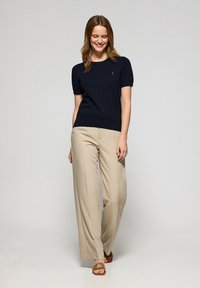 Granatowa koszulka z krótkim rękawem, dzianinowa z prążkowanym dołem i małym czerwonym logo, w zestawie z beżowymi spodniami o szerokich nogawkach i brązowymi klapkami.