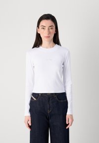 Una giovane donna è in piedi di fronte alla macchina fotografica, indossando un top aderente bianco a maniche lunghe e jeans blue scuri a gamba larga, con un'espressione minima.