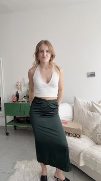 Top halter branco com textura ribada, saia de cetim verde escuro com detalhe franzido, mules pretos e tapete texturizado claro em um interior minimalista.