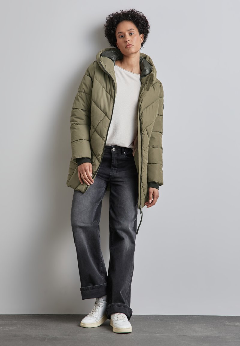 Olivově zelená prošívaná puffer bunda s kapucí, spárovaná s černými širokými džíny a bílými teniskami. Hladký povrch a viditelné švy.