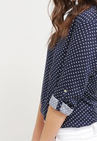 Kvinna iförd en marinblå blus med små vita blommönster och uppvikta ärmar, i kombination med vita byxor, sedd från sidan.