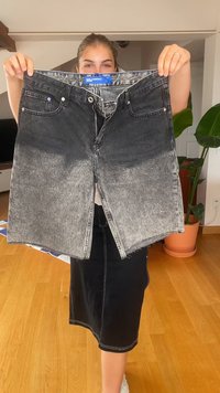 Juodas džinsinis sijonas su išblukusia pilka raštu, su priekiniais užtrauktukais, dviem kišenėmis ir suplyšusiu kraštu. Etiketė matoma ant juosmens.