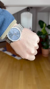 Ασημένιο ρολόι από ανοξείδωτο ατσάλι με μπλε καντράν, ασημένιους δείκτες και τρία χέρια. Φορεμένο σε καρπό με φόντο ξύλινο δάπεδο.