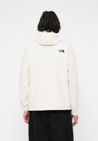 Persona con capelli scuri di lunghezza media che indossa una giacca di pile bianca con cappuccio e il logo The North Face sulla parte superiore della schiena.