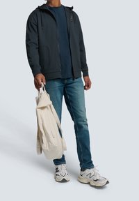 Felpa zip grigia scura, t-shirt blu, jeans azzurri chiari, tiene una borsa di tela beige e sneakers bianche con dettagli neri.
