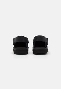 Sorte sandaler med flat såle, med to justerbare stoffstropper og en teksturert fotseng. Bak er åpen for enkel påkledning.