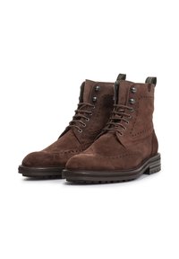 Stivali alla caviglia in suede marrone con punta rotonda, dettaglio wingtip, lacci metallici e suole imbottite. Presentano linguette sul retro per una facile vestibilità.