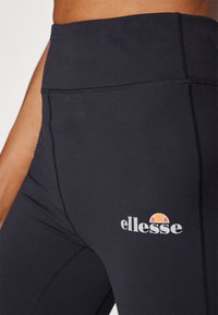 Crne tajice s visokim strukom od rastezljivog materijala, s ravnim šavovima i bijelim "ellesse" logotipom sa narančastim i žutim naglascima.