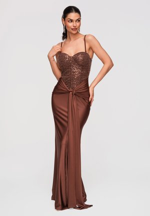 Kobieta w brązowej, cekinowej sukni wieczorowej z gorsetową górą i wiązaniem w talii, pozująca z jedną ręką na ramieniu, a drugą na biodrze.