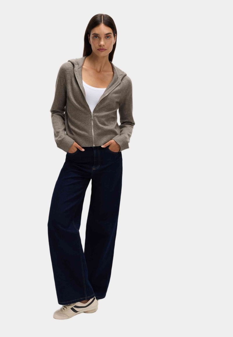 Světle hnědá zipová mikina s texturovaným povrchem, kombinovaná s tmavě modrými kalhotami širokého střihu a béžovými teniskami se černými pruhy.