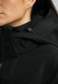 Schwarze Jacke mit hohem Kragen, strukturiertem Stoff, verstellbarem Kordelzug an der Kapuze und einem Reißverschluss vorne. Nahaufnahme des Materials und der Details.