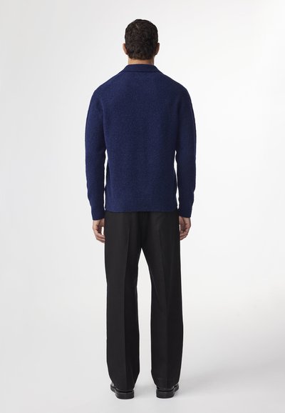 Tummansininen tekstuuri neulepaita, jossa on kaulus, yhdistettynä tummiin suoriin housuihin. Huomio keskittyy paidan rikkaaseen väriin ja materiaaliin.