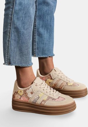 Pieds portant des baskets à plateforme beige avec des sequins floraux et un jean bleu clair court sur fond blanc.